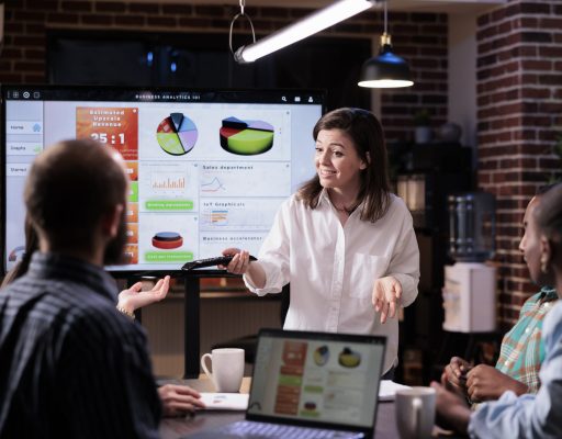 Smiling caucasian woman in startup office doing business presentation on big tv screen with charts in front of team. Confident employee presenting growing sales statistics in late night meeting.